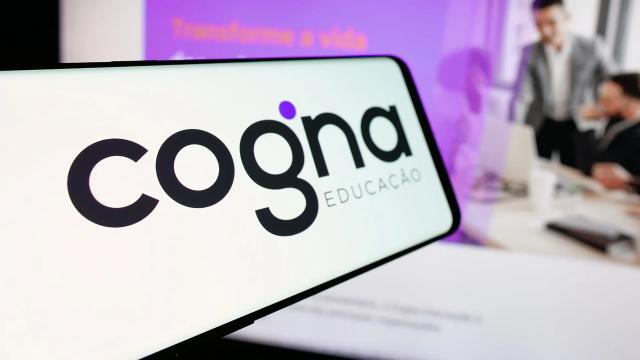 Logotipo da Cogna Educação exibido em tela de celular, com fundo desfocado mostrando ambiente corporativo e materiais institucionais da empresa