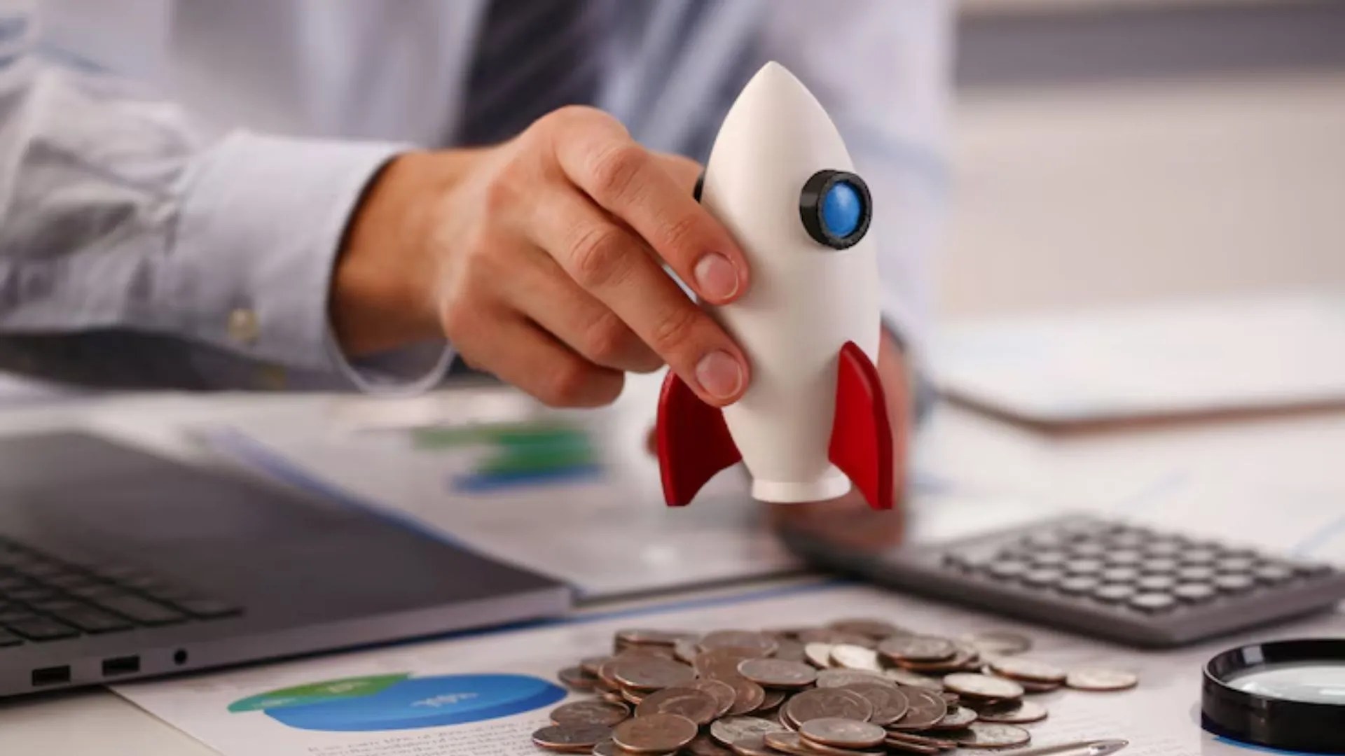 Pessoa segurando um foguete de brinquedo sobre uma mesa com moedas, gráficos e calculadora, simbolizando o início dos investimentos e o crescimento financeiro. Como investir