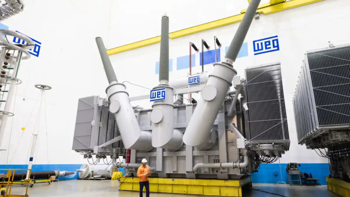 Vista frontal de um grande transformador elétrico industrial da WEG em uma linha de montagem, com um funcionário em uniforme laranja ao lado.