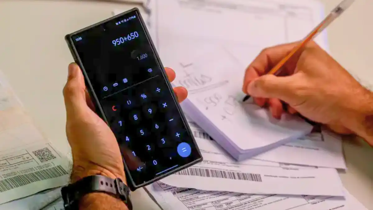 Mão usando o celular como calculadora e outra escrevendo em um bloco de notas.