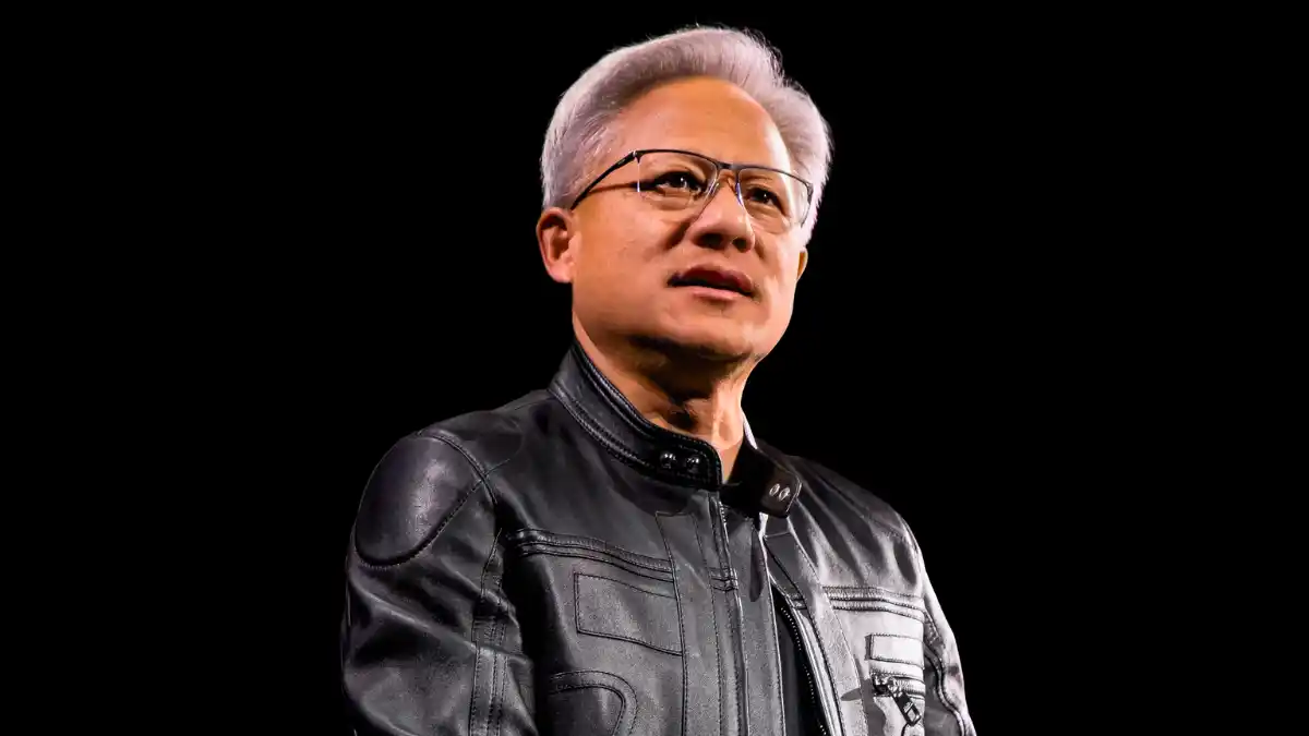 O CEO da Nvidia, Jensen Huang, usando sua jaqueta de couro preta icônica e óculos, em um palco, falando sobre a corrida da Inteligência Artificial (IA).