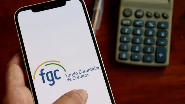 Ilustração do logotipo do FGC em um dispositivo móvel ao lado de cédulas de Real, representando a proteção aos investidores e o início dos pagamentos de garantia no caso Banco Master e Will Financeira em 2026.