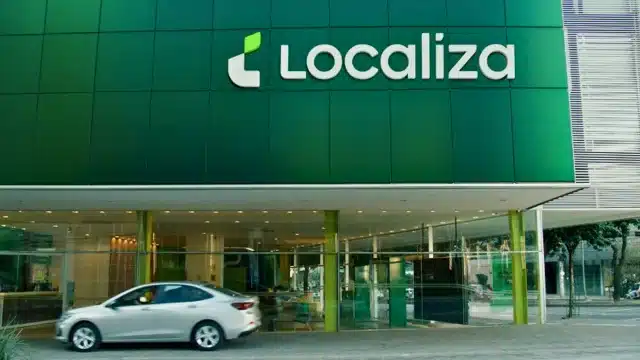 Fachada de uma unidade da Localiza com o logotipo em destaque e um carro prata passando em frente, ilustrando a reabertura da tese de investimento na companhia para 2026.