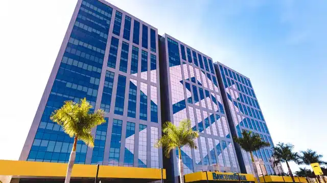 Fachada de um arranha-céu moderno revestido de vidro espelhado com o logotipo gigante do Banco do Brasil refletido, cercado por palmeiras sob um céu limpo, ilustrando a Carteira BB Dividendos.