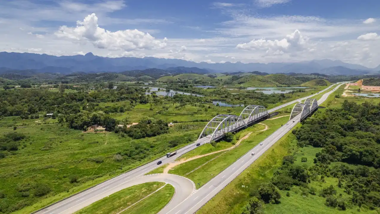 Vista aérea de uma rodovia de pista dupla com pontes em arco metálico atravessando uma área de preservação ambiental com montanhas ao fundo, representando a infraestrutura da Ecorodovias (ECOR3).