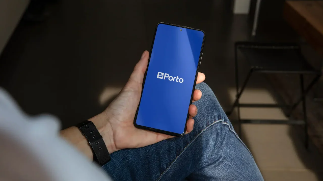 Close-up de uma pessoa segurando um smartphone com a tela azul exibindo o logotipo branco da "Porto". O usuário veste calça jeans e um relógio de pulso preto com pulseira de metal, em um ambiente de luz suave.