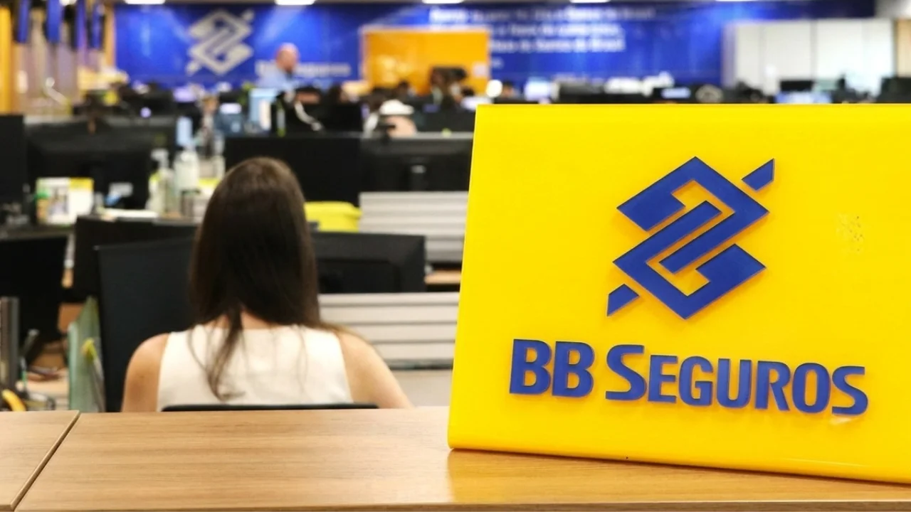 Placa amarela de mesa com o logotipo azul da BB Seguros posicionado em destaque à direita sobre um balcão de madeira. Ao fundo, em ambiente de escritório desfocado, uma funcionária de costas trabalha em sua estação de trabalho diante de monitores de computador, dentro de uma agência ou escritório do Banco do Brasil.