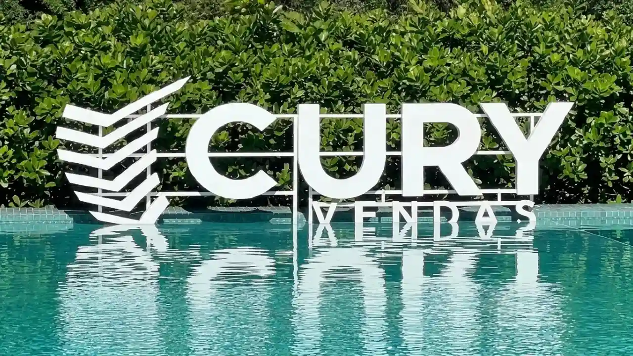 Fachada de um estande de vendas da Cury com o logotipo "CURY VENDAS" em grandes letras brancas instaladas à beira de uma piscina de águas cristalinas. O logotipo e a estrutura metálica branca se refletem perfeitamente na água azul, com uma densa vegetação verde ao fundo. A cena transmite uma sensação de solidez, lazer e sucesso imobiliário.