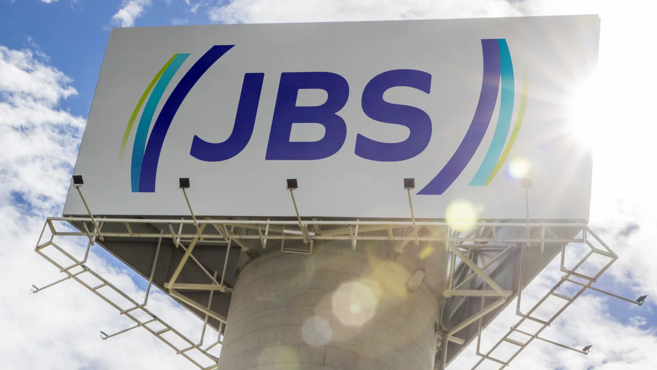 Outdoor branco de grandes dimensões com o logotipo da JBS em azul e detalhes em verde e ciano. A estrutura metálica que sustenta a placa está posicionada contra um céu azul com nuvens esparsas, com o reflexo do sol (lens flare) no canto superior direito.