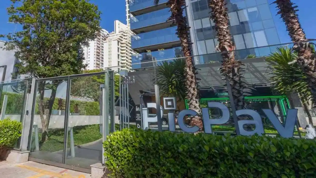 Fotografia em plano aberto e perspectiva diagonal da entrada externa do escritório do PicPay. Em primeiro plano, destaca-se o logotipo da marca em um letreiro tridimensional com letras cinzas texturizadas, instalado logo atrás de uma cerca viva de arbustos verdes e densos. À esquerda, vê-se um portão de segurança de vidro com moldura metálica cinza e árvores frondosas. Ao fundo, ergue-se a estrutura de um edifício comercial moderno com fachada espelhada de vidro azulado, ladeada por duas palmeiras altas sob um céu azul e ensolarado.