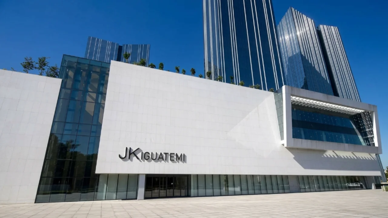 Fotografia arquitetônica em plano aberto e ângulo contra-plongé suave da fachada externa do Shopping JK Iguatemi em um dia de céu azul limpo. O edifício exibe uma enorme estrutura minimalista de placas de concreto branco fosco com a inscrição metálica em alto-relevo "JK IGUATEMI" centralizada. Logo acima e ao fundo, erguem-se modernos arranha-céus corporativos espelhados com faixas verticais prateadas que refletem a luz solar intensa. O piso em primeiro plano é composto por uma esplanada de blocos de granito claro totalmente desimpedida.