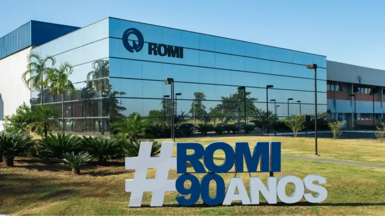 Fotografia em plano aberto e ângulo lateral da fachada frontal da sede da Romi em um dia ensolarado. O moderno edifício industrial possui uma grande extensão de vidro espelhado que reflete o céu azul claro, exibindo no topo esquerdo o logotipo circular da marca e o nome "ROMI" em letras pretas em alto-relevo. Em primeiro plano, sobre um gramado verde aparado com arbustos e palmeiras de jardim, destaca-se um letreiro tridimensional com uma hashtag branca e letras e números em azul escuro formando a inscrição "#ROMI 90 ANOS".