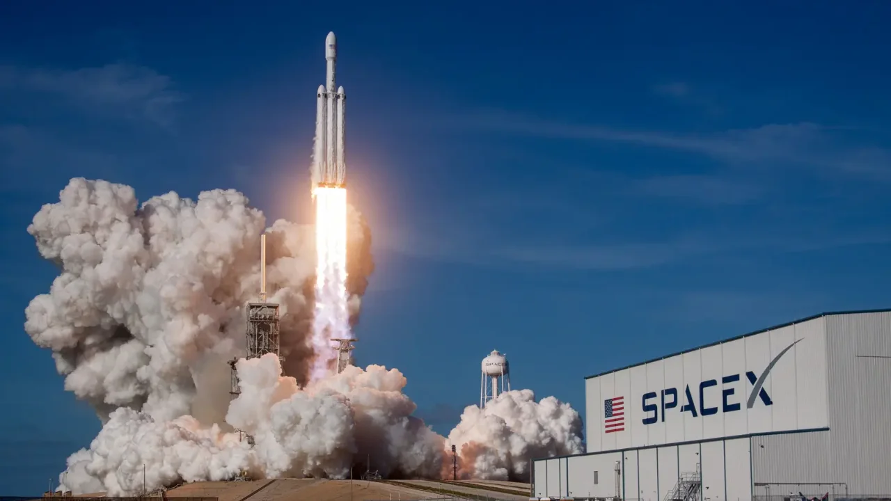 Fotografia em plano aberto capturando o momento exato do lançamento de um imponente foguete espacial branco de múltiplos propulsores decolando verticalmente de sua base. O veículo deixa um rastro de chamas alaranjadas e luzes intensas, gerando uma massiva e densa nuvem de fumaça branca e cinza que se expande pelo solo. À direita, em primeiro plano, destaca-se um grande galpão industrial e hangar branco com o logotipo da marca "SPACEX" e a bandeira dos Estados Unidos estampados na lateral. O cenário de fundo exibe um céu azul claro e limpo.