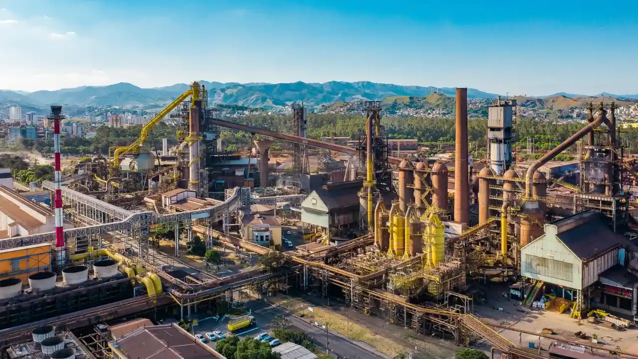 Fotografia aérea panorâmica em grande plano geral do complexo industrial e siderúrgico da Usiminas sob um céu azul limpo. A imagem revela uma vasta infraestrutura de engenharia pesada, composta por altos-fornos cilíndricos em tons de marrom e ferrugem, estruturas metálicas complexas, chaminés elevadas (uma delas listrada em vermelho e branco na lateral esquerda) e longas esteiras transportadoras protegidas por coberturas amarelas. No pátio térreo da usina, observam-se vias internas pavimentadas com pequenos veículos estacionados. Ao fundo, o cenário industrial é emoldurado por colinas e montanhas verdes, dividindo espaço com áreas residenciais urbanas da região.