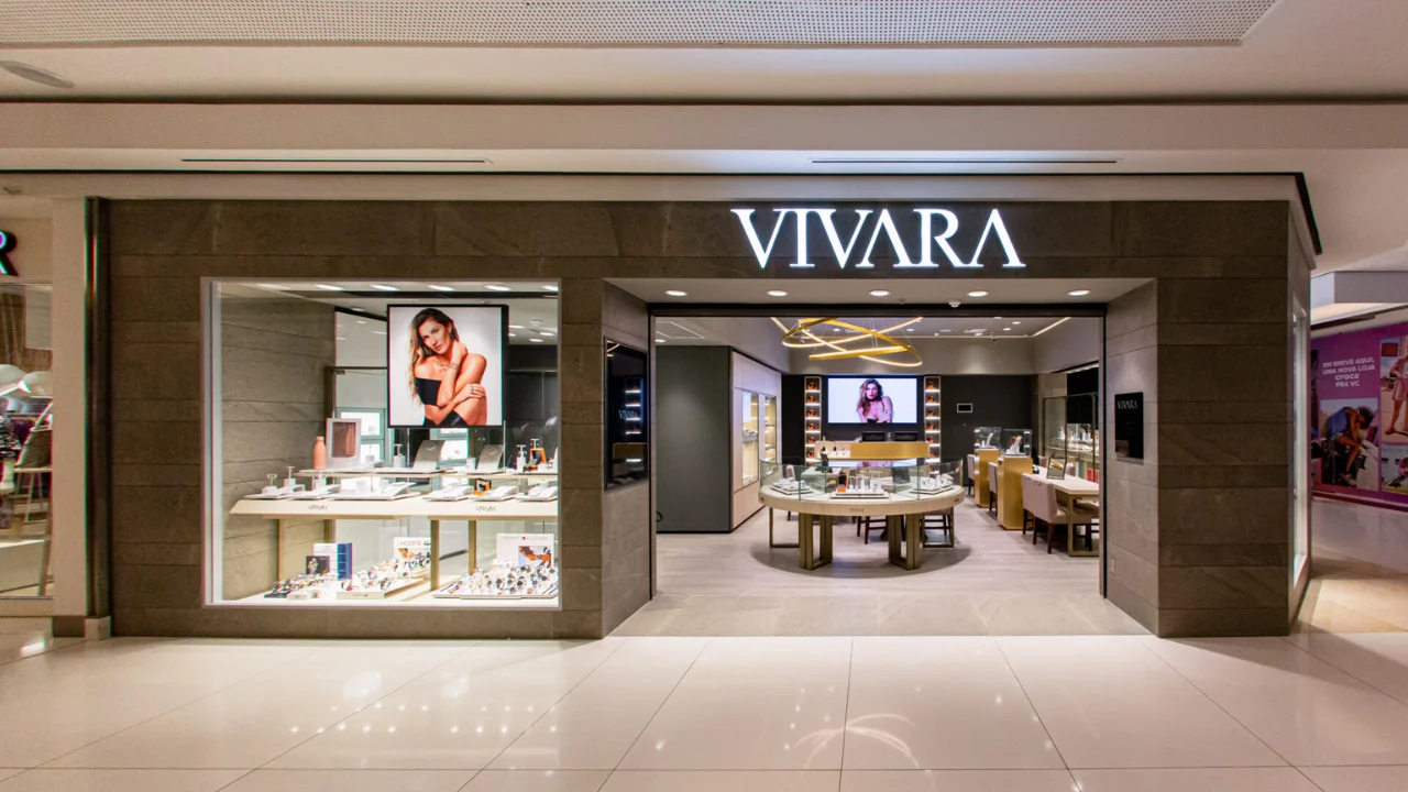 Fotografia frontal da fachada de uma sofisticada loja de joias Vivara em um shopping center. O pórtico externo é revestido em pedras em tons de cinza e marrom-claro, ostentando um grande letreiro luminoso com a palavra "VIVARA" em letras maiúsculas brancas no topo central. Na vitrine de vidro à esquerda, destaca-se um grande cartaz iluminado da modelo Gisele Bündchen posando com joias, sobre expositores com relógios e acessórios. A entrada ampla revela o interior iluminado da loja, com balcões circulares e retangulares de vidro para exibição de produtos, cadeiras estofadas e iluminação embutida sobre o piso claro e brilhante do corredor.