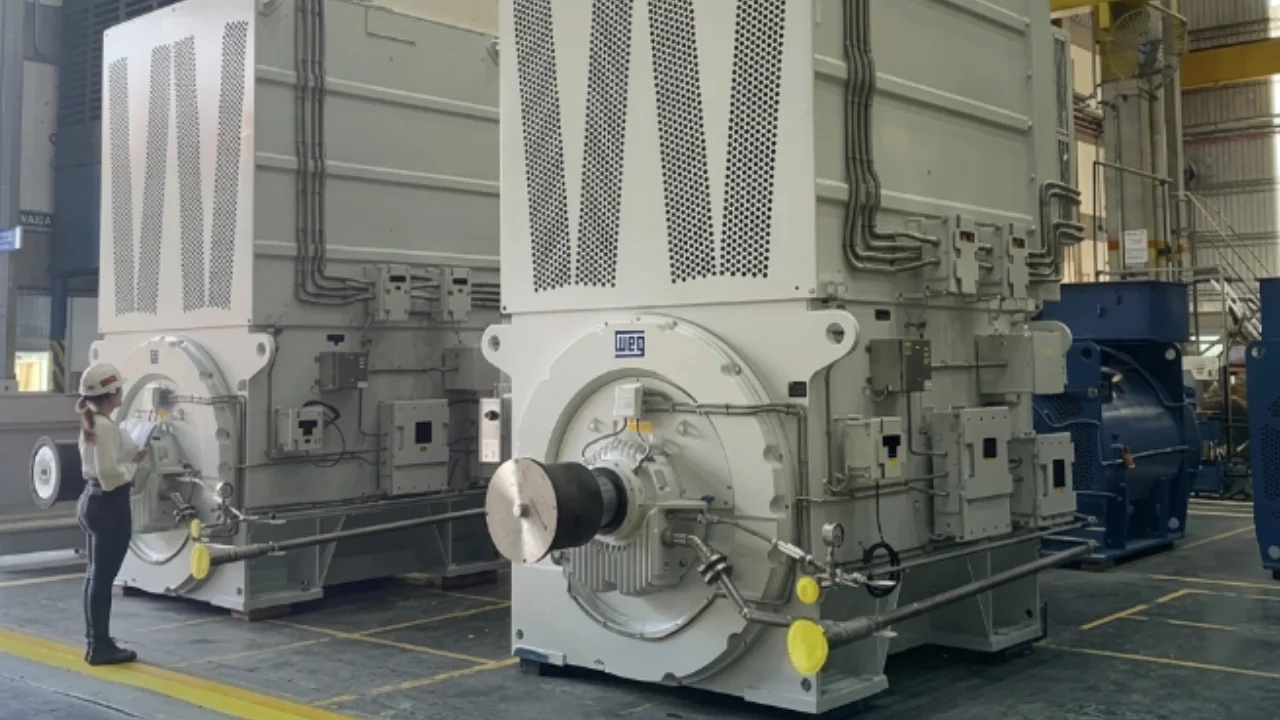 Fotografia em plano médio no chão de fábrica da WEG. Em primeiro plano à esquerda, uma inspetora com capacete branco de segurança, calça escura e camisa clara segura uma prancheta enquanto analisa um gigantesco gerador/motor industrial de cor branca. Ao centro, destaca-se um segundo gerador de grandes proporções no mesmo tom branco com a marca "WEG" em azul gravada na estrutura circular frontal. À direita, ao fundo, observa-se outro equipamento de grande porte na cor azul-marinho no galpão industrial iluminado.