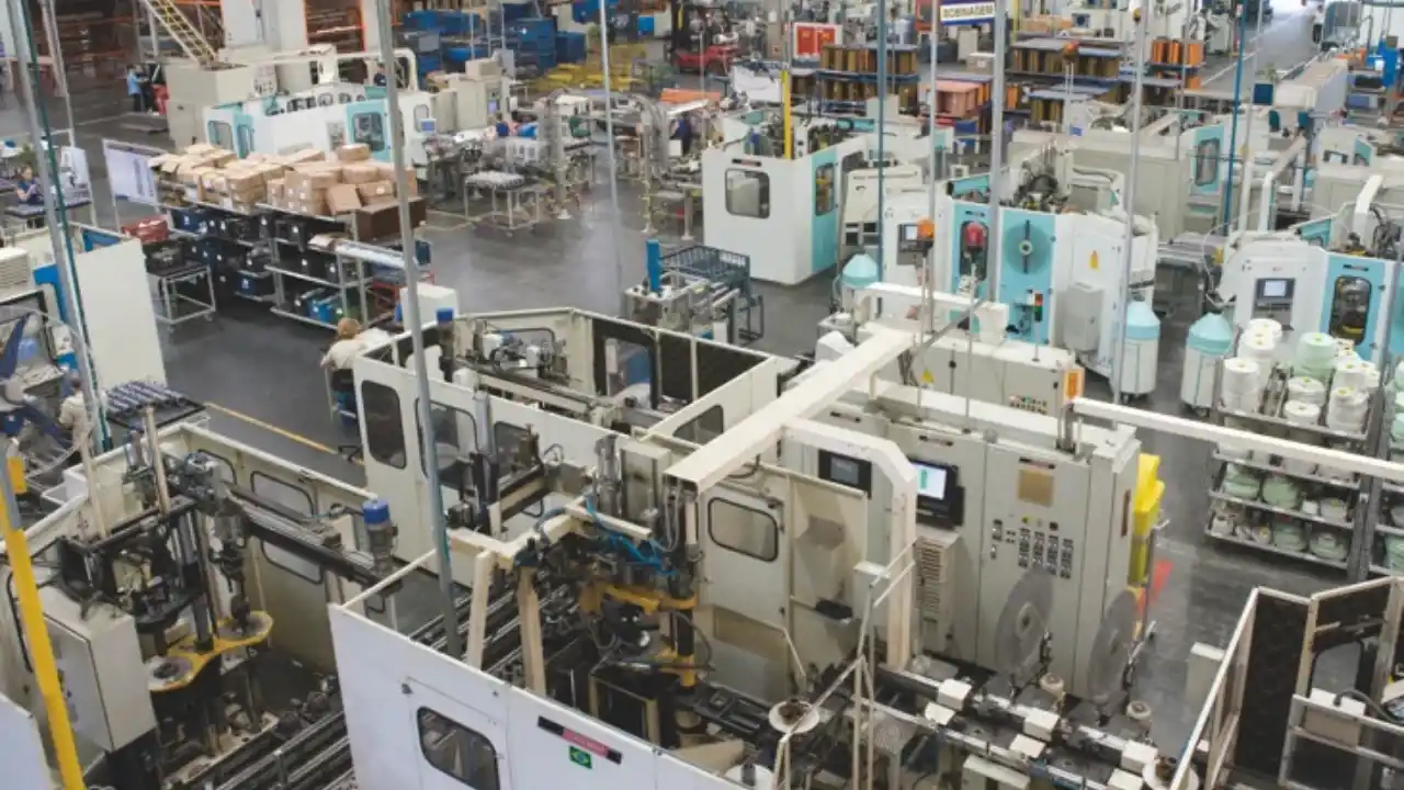 Fotografia industrial em plano geral e ângulo elevado do pátio de produção de uma fábrica da WEG. O amplo galpão exibe diversas células de trabalho automatizadas, maquinários industriais de grande porte em tons de branco e detalhes em azul-claro, esteiras transportadoras e bancadas de montagem. À esquerda, veem-se prateleiras de aço com caixas de papelão organizadas e componentes eletrônicos. No topo de uma das máquinas centrais, há uma placa de identificação com o texto "BOBINAGEM". Operários de uniforme escuro podem ser observados operando discretamente os sistemas ao longo do piso industrial cinza refletivo.