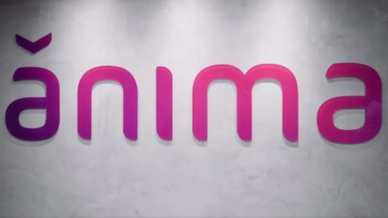 Fotografia em plano frontal do logotipo corporativo em alto-relevo da "ânima" fixado em uma parede de concreto queimado em tom cinza-claro. As letras arredondadas em acrílico apresentam um gradiente de cores que varia do roxo no acento circunflexo invertido e na letra "â" inicial até o rosa-choque brilhante na letra "a" final. Três pontos de iluminação dicroica no teto direcionam luz suave sobre a marca.