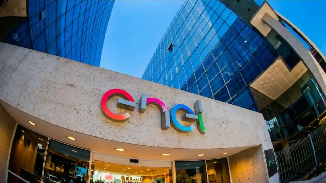 Fotografia corporativa em contraponto (ângulo baixo) da sede institucional da Enel São Paulo. Em primeiro plano, destaca-se um pórtico de fachada em pedra clara com o logotipo luminoso "enel" em degradê de vermelho, rosa, azul e verde. Ao fundo e acima, ergue-se um moderno edifício comercial com fachada de vidro azul reflexivo sob céu aberto.