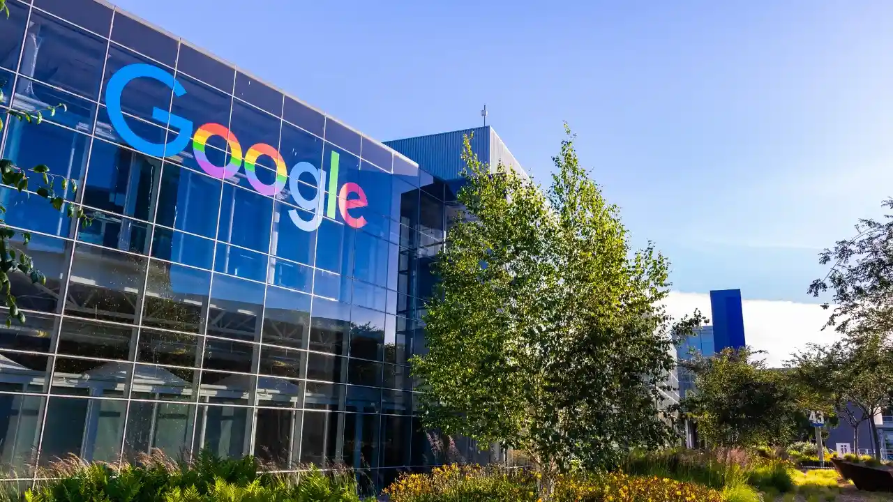 Fotografia em ângulo contraponto da moderna fachada envidraçada do edifício corporativo do Google. Na altura dos andares superiores da estrutura de vidro, destaca-se o logotipo oficial do Google estilizado em cores vibrantes do arco-íris. Em primeiro plano, veem-se canteiros ornamentais com arbustos verdes e flores amarelas, além de árvores que moldam a composição ao lado sob céu azul de dia ensolarado.