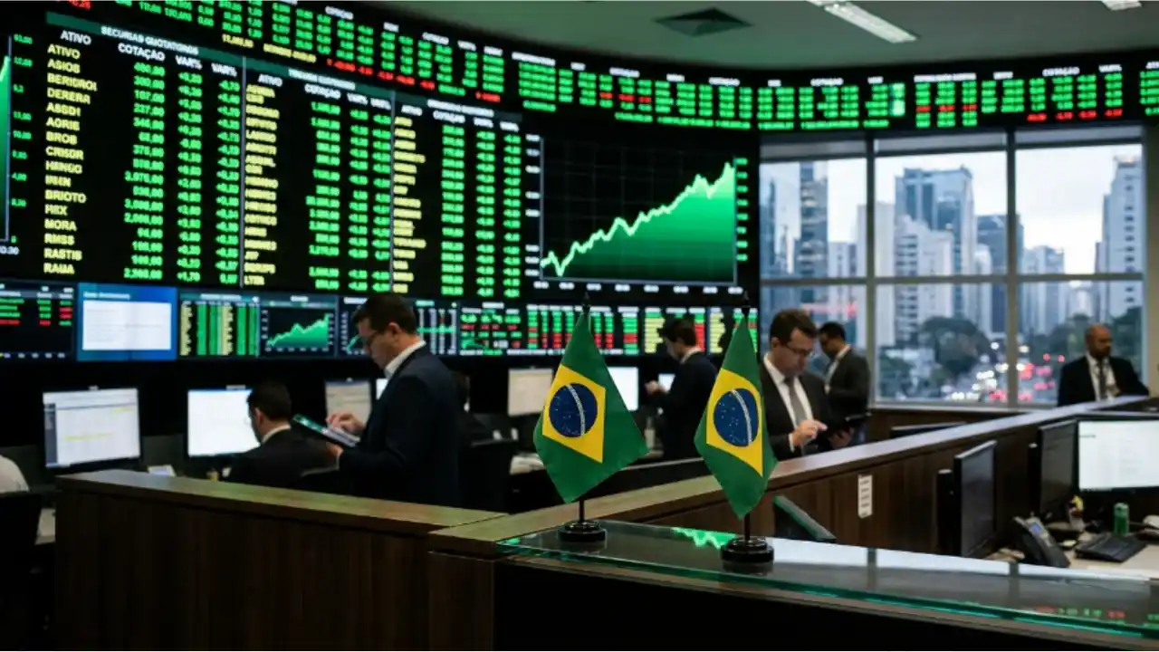 Vista interna de uma sala de operações financeiras (trading floor) com profissionais em trajes formais trabalhando em monitores e telefones. Ao fundo, um imponente painel eletrônico de alta definição exibe uma grande grade de cotações em verde e um gráfico de linha verde com inclinação ascendente acelerada. Em primeiro plano, sobre a divisória de madeira da bancada, estão posicionadas duas pequenas bandeiras do Brasil, com amplas janelas ao fundo mostrando a vista de edifícios corporativos.