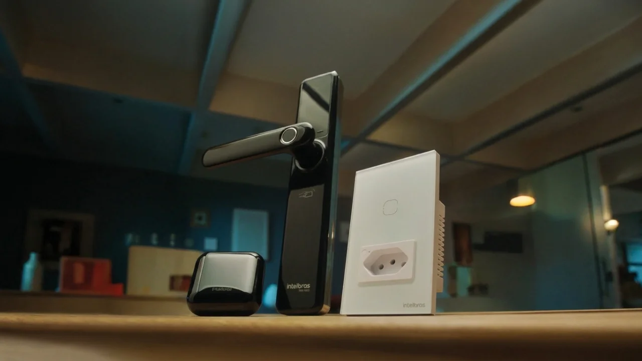 Fotografia em plano de detalhe sobre uma bancada de madeira exibindo produtos da linha de inteligência e automação residencial da Intelbras. Ao centro, destaca-se uma fechadura digital vertical de cor preta com leitor biométrico de impressão digital e a marca "intelbras" impressa na base. À direita, um painel de interruptor e tomada inteligente branco com acabamento em vidro ("intelbras"). À esquerda, um mini hub de conectividade preto sob iluminação ambiente aconchegante.
