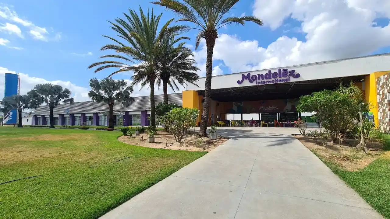 Fotografia em plano amplo da fachada externa de um centro corporativo e fabril da Mondelēz International sob dia ensolarado. Em primeiro plano, observa-se um amplo gramado verde com caminho de concreto cinza que conduz à entrada principal. No alto da estrutura da cobertura, destaca-se a marca em letras roxas "Mondelēz International", emoldurada por palmeiras ornamentais e céu azul com poucas nuvens.