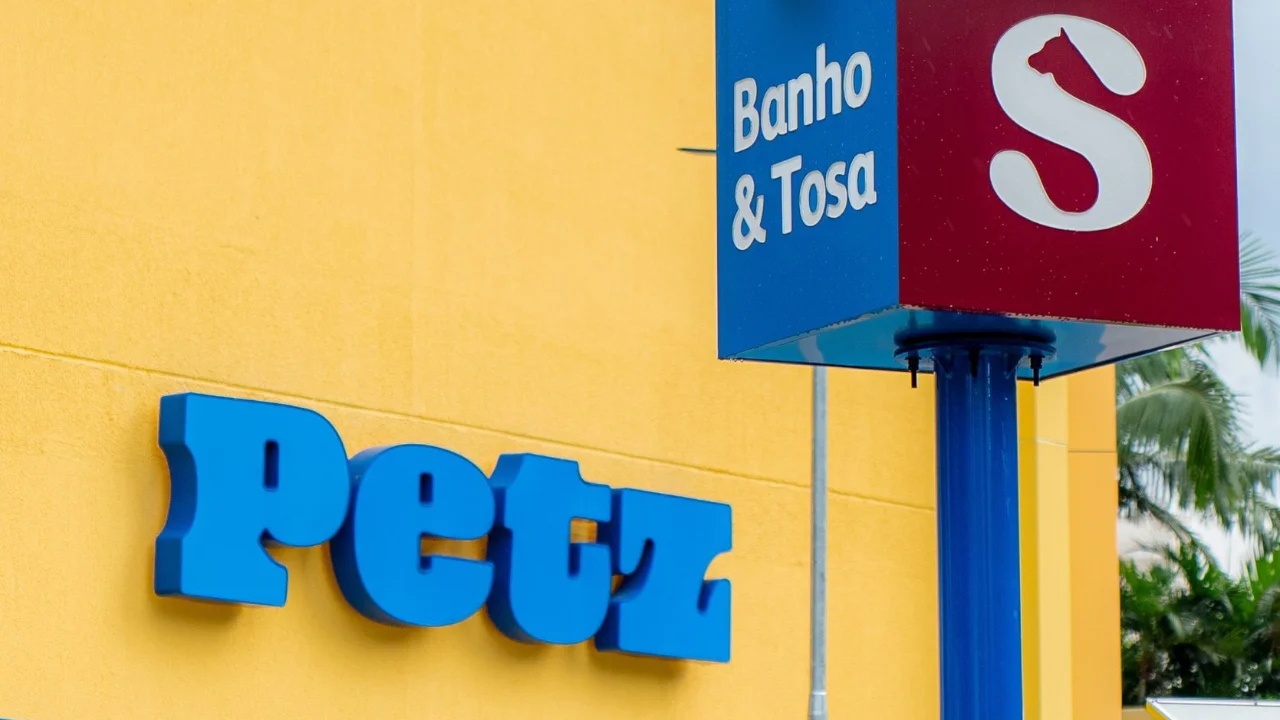 Fotografia em plano médio da fachada exterior de uma megaloja da rede Petz. À esquerda, sobre uma parede pintada em amarelo vivo, destaca-se o logotipo "Petz" com letras estilizadas em alto-relevo na cor azul. À direita, sobressai-se a estrutura de um totem metálico azul sustentando uma placa cúbica com a indicação "Banho & Tosa" em letras brancas sobre fundo azul e o símbolo corporativo com a silhueta de um cão em fundo vermelho. Ao fundo, folhagens de palmeiras verdes aparecem sob iluminação natural sob céu claro.