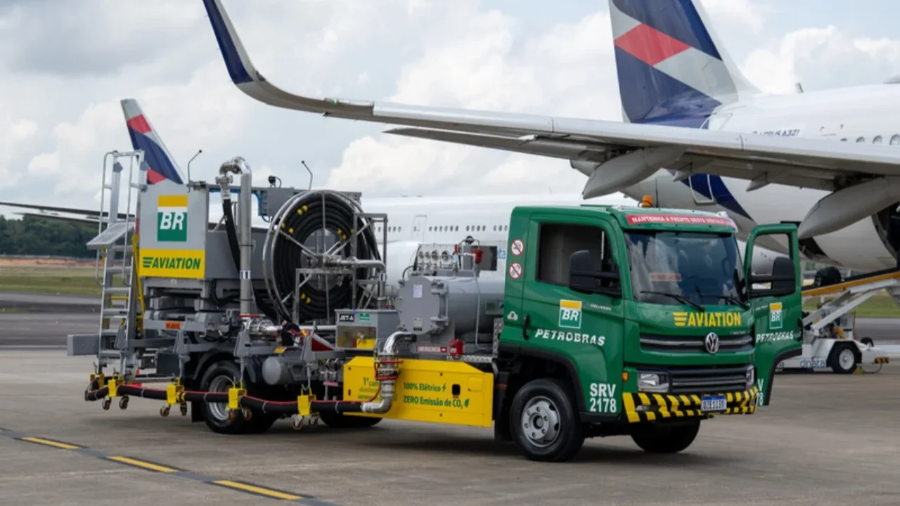 Fotografia em plano inteiro de um caminhão-tanque elétrico de abastecimento verde e amarelo da BR Aviation (Petrobras), modelo Volkswagen Delivery, estacionado na pista de concreto de um aeroporto sob céu nublado. O veículo exibe os logotipos "BR" e as inscrições "BR PETROBRAS" e "BR AVIATION", além de um adesivo lateral amarelo com os dizeres "1º caminhão de QAV 100% Elétrico - ZERO Emissão de CO2". Na parte traseira, o caminhão possui um complexo sistema de mangueiras pretas enroladas em uma bobina metálica e tubulações para bombeamento de combustível. Ao fundo, a asa e parte da fuselagem traseira de um avião comercial de passageiros com pintura branca e listras azuis e vermelhas na cauda estão posicionadas para receber o serviço de solo.
