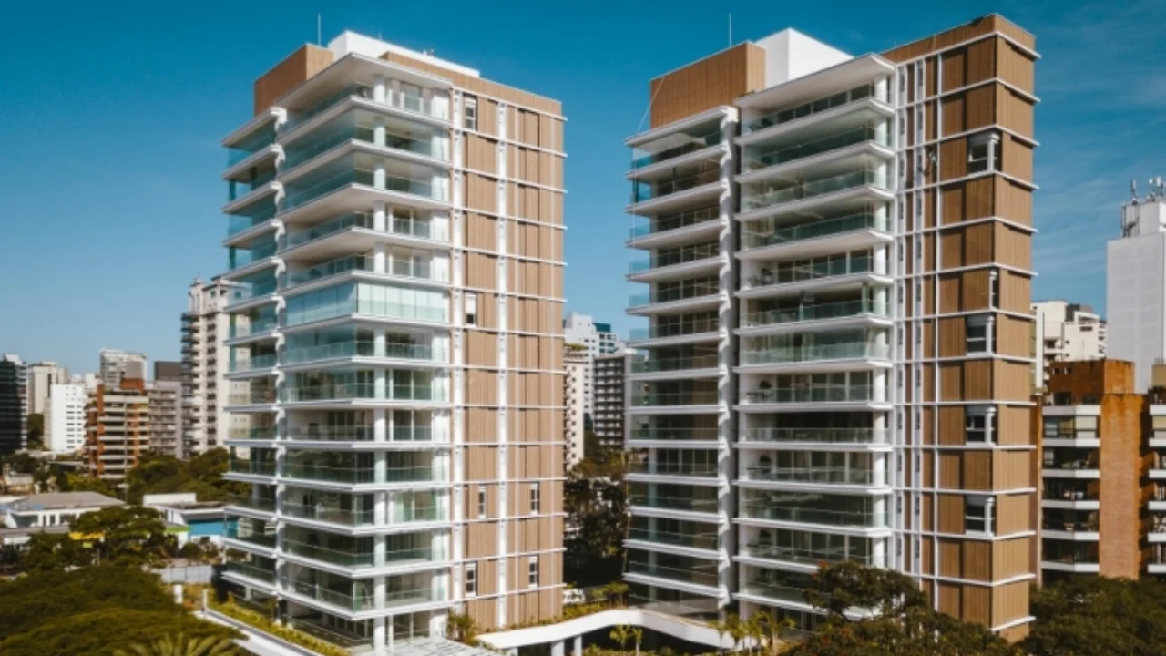 Fotografia em ângulo frontal e elevação vertical de duas imponentes torres residenciais contemporâneas da Trisul. Os edifícios contam com varandas envidraçadas com guarda-corpos em vidro, colunas de revestimento amadeirado e detalhes em tons de bege e branco. Ao centro, um corredor elevado com cobertura curva conecta as estruturas na base. Ao fundo, avista-se a copa de árvores verdes e a paisagem urbana de edifícios vizinhos sob um céu azul sem nuvens.