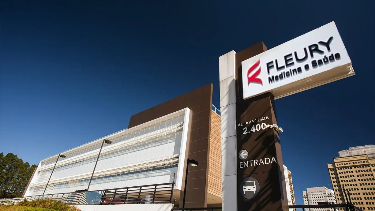 Fachada moderna e espelhada de unidade do Fleury Medicina e Saúde em ângulo inferior sob céu azul, com totem marrom e branco contendo o logotipo da marca e sinalização de entrada na Alameda Araguaia 2400.