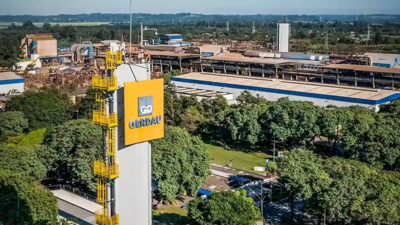 Vista panorâmica aérea de uma usina siderúrgica da Gerdau, com torre vertical exibindo a logomarca amarela da empresa em primeiro plano, cercada por áreas verdes e galpões industriais.