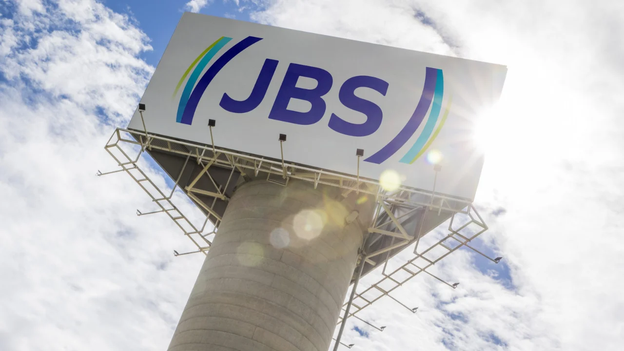 Torre de concreto sustentando um grande painel publicitário com a logomarca azul e verde da JBS contra o céu parcialmente nublado e ensolarado.
