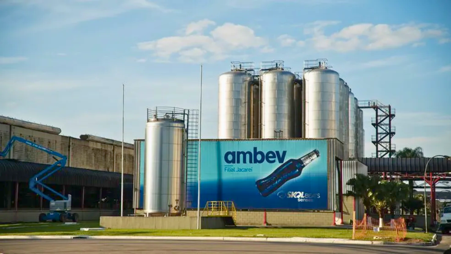 Grande instalação industrial com vários tanques de armazenamento altos e cilíndricos. Na frente desses tanques, há um outdoor com o logotipo da “Ambev” ao lado de uma imagem de uma garrafa azul.