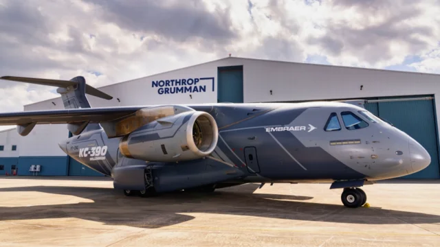 Aeronave militar cargueira KC-390 Millennium da Embraer estacionada em um pátio, com um hangar da Northrop Grumman ao fundo, simbolizando a parceria estratégica Embraer Northrop.