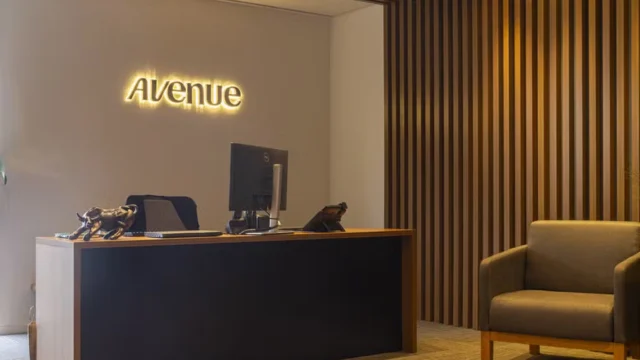 Recepção moderna de um escritório da Avenue, com balcão de atendimento preto, poltrona cinza e logotipo iluminado na parede ao fundo, representando a expansão de Avenue produtos.