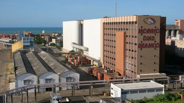 Vista aérea do complexo industrial Moinho Dias Branco, apresentando grandes silos brancos, galpões logísticos e o prédio administrativo com o logotipo da empresa em frente ao mar, representando a m. dias.