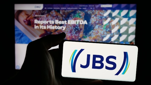 Logo da JBS em primeiro plano, em tela de celular à frente de uma tela de computador