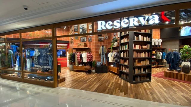 Fachada moderna de uma loja da marca Reserva, integrante do grupo Azzas 4t25, em um shopping, com interior iluminado, estantes de livros e araras de roupas em tons amadeirados.