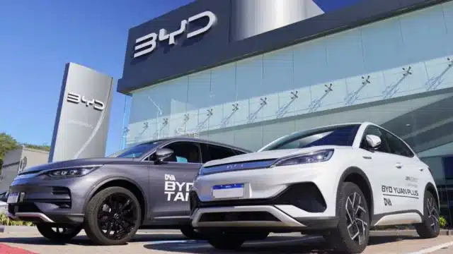 Dois carros elétricos modernos, um cinza modelo BYD TAN e um branco modelo BYD YUAN PLUS, estacionados em frente a uma concessionária envidraçada da BYD sob céu azul, ilustrando o cenário de montadoras chinesas impacto locadoras.