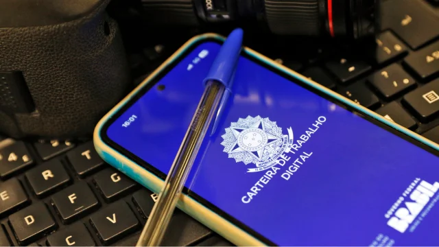 Close-up de um smartphone com a tela ligada exibindo o aplicativo da Carteira de Trabalho Digital do Governo Federal do Brasil. O fundo da tela é azul escuro com o Brasão da República em branco e o texto "CARTEIRA DE TRABALHO DIGITAL". Sobre o celular, há uma caneta esferográfica azul transparente. O aparelho está apoiado sobre um teclado de computador preto e há uma câmera fotográfica profissional desfocada ao fundo.