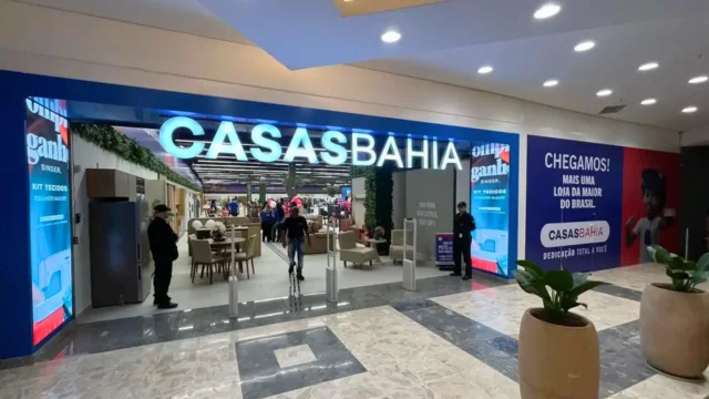 Fachada de uma loja da Casas Bahia em um shopping, com o letreiro iluminado em azul e branco, exibindo o interior com móveis e eletrodomésticos, representando as ações Casas Bahia (BHIA3).