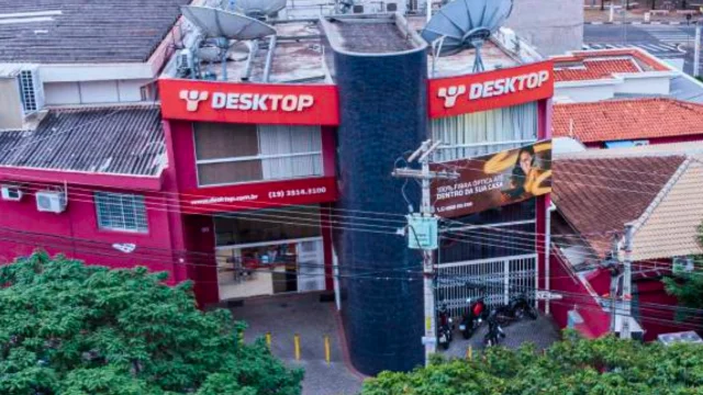 Vista aérea da fachada de uma unidade operacional da Desktop (provedora de internet), com letreiros vermelhos e antenas parabólicas no telhado, ilustrando a estrutura por trás dos resultados da desktop 4t25.