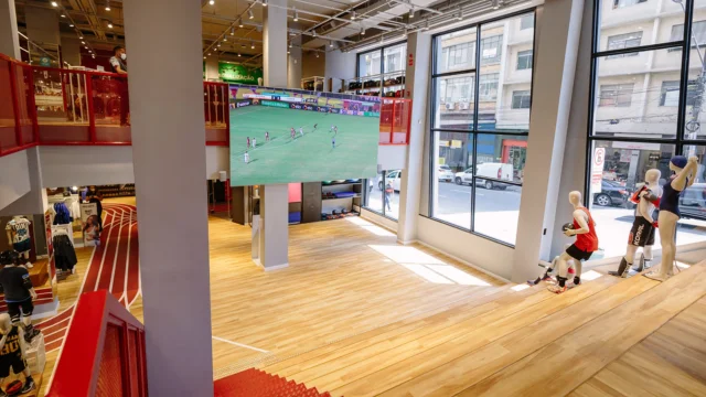 Vista interna de uma loja de esportes do Grupo SBF, com amplo piso de madeira, manequins esportivos junto a janelas de vidro e um grande telão exibindo uma partida de futebol, ilustrando a estratégia do Grupo SBF ataque.