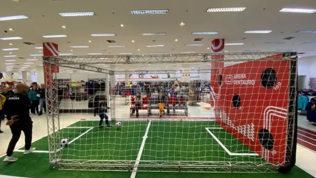 Vista aérea de um gigantesco centro de distribuição logística do Grupo SBF, apresentando um prédio industrial branco com telhado extenso e dezenas de docas para caminhões, ilustrando a preparação para a Grupo SBF Copa do Mundo.
