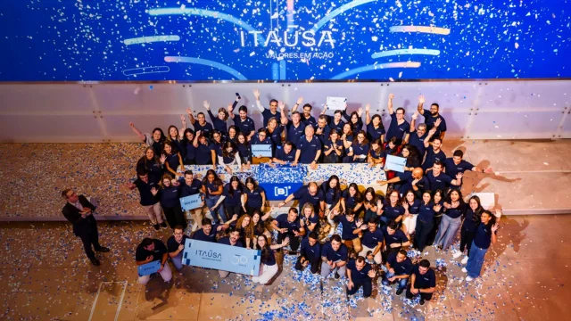 Grande grupo de colaboradores da Itaúsa vestindo camisas polo azuis, reunidos em um palco sob chuva de confetes azuis e brancos, celebrando os 50 anos da companhia em um evento com a marca B3 ao centro, ilustrando o valor da gestão e o desconto de holding itaúsa.