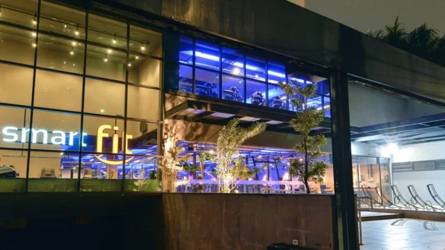 Fachada externa noturna de uma academia da rede Smart Fit com grandes janelas de vidro iluminadas, exibindo o logotipo amarelo e áreas de treino internas, ilustrando a expansão da Smart Fit 4t25