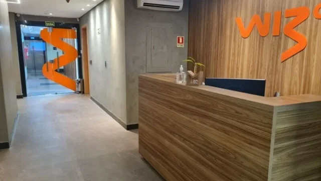 Escritório moderno da Wiz Co com estações de trabalho brancas, cadeiras pretas e o logotipo da empresa em laranja e amarelo em uma parede de destaque, representando a sede para análise da Wiz WIZC3.