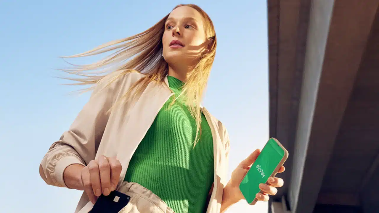 Mulher loira de casaco bege e blusa verde caminhando ao ar livre, segurando um smartphone com o aplicativo do PicPay aberto e retirando um cartão de crédito preto do bolso.