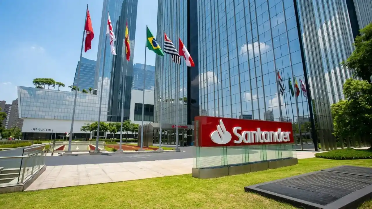 Vista de baixo para cima do topo de um edifício comercial moderno com o logotipo vermelho do Banco Santander no topo. O prédio possui fachada de vidro espelhado que reflete um céu azul claro. No canto inferior, aparecem as copas de árvores verdes e parte de uma estrutura metálica.